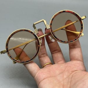 May include: Round eyeglasses featuring a gold-tone metal frame with a tortoiseshell pattern on the rims. The bridge has a distinct geometric, elevated square shape connecting the two circular lenses, which have a light brown tint.