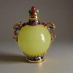 May include: A decorative, yellow-green glass bottle with a gold-coloured metal base and accents. The bottle features a round shape, ornate dragon handles, and a jeweled stopper with red, green, and blue stones. The bottle is approximately 10 cm tall.