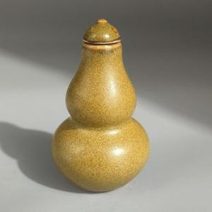 May include: A gourd-shaped ceramic vessel with a speckled olive-green glaze and a matching removable lid. The container features a rounded base and a narrower upper section, creating a distinct hourglass silhouette with a smooth, glossy finish.