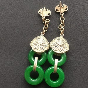 May include: A pair of dangle earrings featuring silver-toned metal tops with a decorative embossed pattern, connected by short chains to two stacked circular green jade-like stone rings. The earrings have a metallic post backing for attachment.