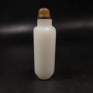 May include: A cylindrical white glass snuff bottle with a smooth, opaque finish. The bottle features a narrow neck topped with a metallic, textured collar and a rounded, amber-colored stopper that sits securely at the very top of the vessel.