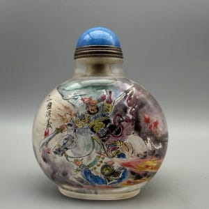 May include: A round, translucent snuff bottle featuring a hand-painted scene of a warrior on horseback wielding a sword. The bottle is topped with a smooth, bright blue spherical stopper and includes black Chinese calligraphy characters on the side.