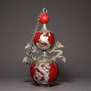 May include: A decorative red and silver bottle with a chain. The bottle has a round shape with silver dragon designs and a red sphere at the top. The bottle is an antique and has a unique design.