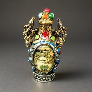 May include: A decorative snuff bottle featuring a metallic gold and silver finish with ornate dragon-shaped handles. The center displays an oval landscape painting, accented by colorful inlaid stones in red, orange, green, and blue on the body and lid.