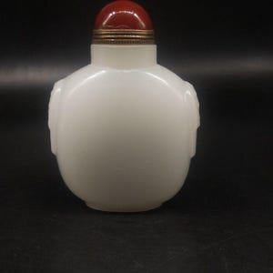 May include: A small, opaque white snuff bottle with a rounded, flattened body and subtle carved side details. The bottle features a smooth, milky finish and is topped with a rounded, deep red cap secured by a metallic, ribbed collar at the neck.