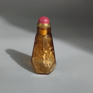 May include: A small, amber-colored glass snuff bottle with a faceted, tapered body containing gold-colored flecks. The bottle features a brass-colored metal neck and a rounded, textured pink stopper at the top, creating a distinct contrast.