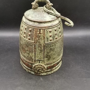 Puede incluir: Una campana cilíndrica de bronce con un acabado de pátina verde desgastada. La campana presenta un asa superior con forma de dos criaturas estilizadas, una serie de tachuelas circulares en relieve, divisores de paneles y un emblema circular.
