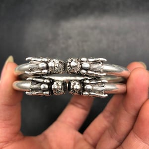 May include: Two silver-colored dragon head bracelets. Each bracelet features a detailed dragon head design at the ends. The bracelets are held together, showcasing the intricate craftsmanship and metallic sheen.