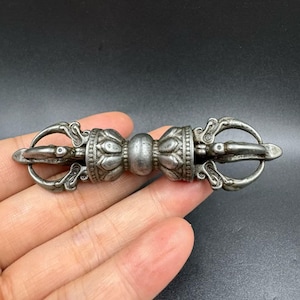 Puede incluir: Un objeto ritual vajra de metal en tono plateado con un agarre central esférico flanqueado por diseños simétricos de pétalos de loto y puntas curvas en ambos extremos. El artículo tiene un acabado mate desgastado con detalles en relieve.