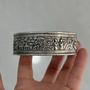 Puede incluir: Un brazalete plateado grabado con un diseño en relieve detallado. El diseño incluye figuras, nubes y elementos arquitectónicos. El brazalete se sostiene en una mano, mostrando la intrincada artesanía y la forma general.