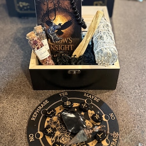 May include: A wooden box set containing a deck of cards with a raven illustration, a black beaded necklace, a raven pendant, two small glass bottles, a bundle of sage, and a wooden stick. A black and gold divination board is in the foreground.