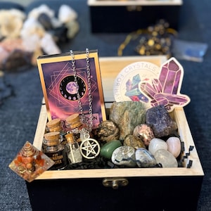 May include: A black wooden box filled with crystals, small glass bottles, a necklace with a pentagram pendant, and a deck of cards. The box also contains a sticker with the words "Crystals for Happy".