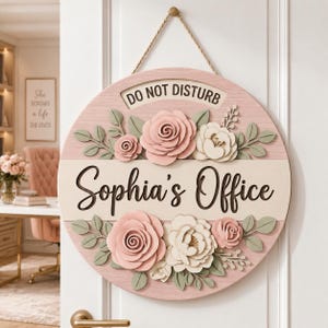 May include: A round wooden door sign featuring a light pink background with a horizontal cream-colored stripe. The text reads "DO NOT DISTURB" and "Sophia's Office" in dark brown script. It is decorated with layered pink and cream paper flowers.