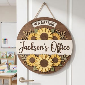 May include: A circular wooden door sign featuring a dark brown wood grain background, yellow sunflowers with green leaves, and a cream-colored horizontal center strip with the text Jackson's Office and a top tab reading IN A MEETING.