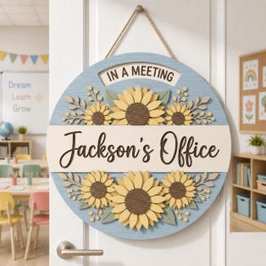May include: A round wooden door sign with a light blue background, featuring yellow sunflowers and green leaves. A central white horizontal band displays the text "Jackson's Office" in black, with "IN A MEETING" printed above it. It hangs by a rope.