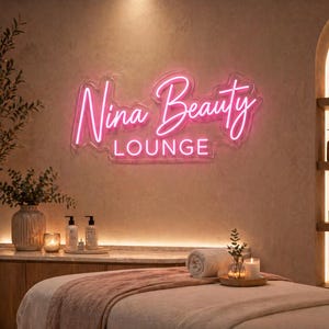 May include: A spa interior featuring a pink neon sign that reads "Nina Beauty Lounge." The room has a massage table with a pink blanket, a wooden shelf with beauty products, and decorative plants. The overall ambiance is warm and inviting.