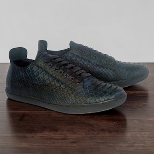 May include: A pair of low-top sneakers featuring a dark blue, textured, scale-like pattern across the upper. The shoes include black laces, a rounded toe, and a matching dark blue flat rubber sole, with a prominent pull tab at the heel collar.