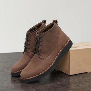May include: A pair of brown leather ankle boots with a lace-up design. The boots have a dark brown sole and a stitched detail around the base. A cardboard box is in the background.