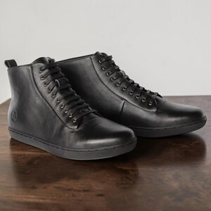 May include: A pair of black leather high-top sneakers featuring a smooth finish, matching black laces, silver-toned metal eyelets, and a flat black rubber sole. A small circular logo is embossed on the outer side of the heel area of each shoe.