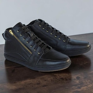 May include: A pair of black high-top sneakers featuring a side zipper with gold-toned hardware, black laces, and matching black eyelets. The shoes have a smooth finish, a rounded toe, and a flat black rubber sole, providing a clean and uniform look.