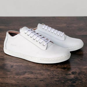 May include: A pair of low-top sneakers in solid white with a smooth finish, featuring white laces, silver-toned eyelets, and a textured white rubber sole. The side of the shoe displays a subtle embossed logo, while the interior lining is a light tan.