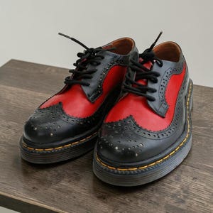 May include: A pair of black and red leather brogue shoes with black laces. The shoes have a thick black sole with yellow stitching. The red leather is on the sides and the black leather is on the toe and heel.