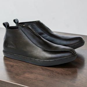 May include: A pair of black ankle boots featuring a smooth leather-like finish, a rounded toe, and a pull tab at the heel. The boots have a side zipper closure, a textured black rubber sole, and the word Atmel embossed on the outer side panel.