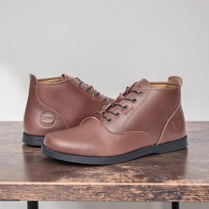 May include: A pair of brown leather ankle boots with black soles and laces. The boots have a round logo on the side and are displayed on a wooden surface. The boots are a medium brown color.