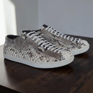 May include: A pair of low-top sneakers featuring a grey and white snakeskin-patterned upper, white laces, and a solid white rubber sole. The textured material covers the entire exterior of the footwear, providing a consistent scale-like appearance.