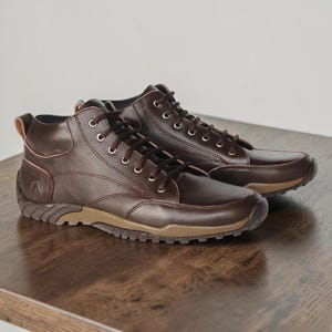 May include: A pair of dark brown leather ankle boots featuring dark brown laces, silver-toned metal eyelets, a tan rubber sole with a textured tread pattern, and a small embossed circular logo on the outer heel area of each shoe.