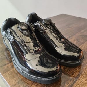 May include: Pair of glossy black shoes with a unique closure system. The shoes have a shiny, patent leather-like finish and a round dial on the top for adjusting the fit. The soles are black and the shoes are sitting on a wooden surface.