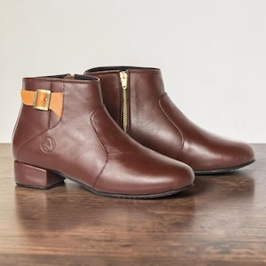 May include: A pair of dark brown ankle boots featuring a low block heel, a side zipper with a gold-tone pull, and a decorative tan strap across the outer ankle secured by a gold-tone buckle. A circular logo is embossed on the side of the boot.