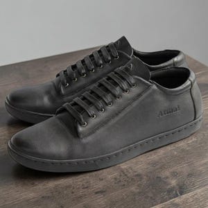 May include: A pair of black leather sneakers featuring a low-top silhouette, matching black laces, and a rounded toe. The side of the shoe displays the embossed text "Atmai" near the heel, while a small logo is visible on the tongue.