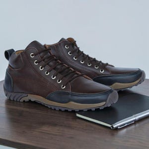 May include: A pair of dark brown leather high-top sneakers featuring black leather toe caps and heel overlays. The shoes have silver metal eyelets, dark brown laces, a pull tab at the heel, and a tan rubber sole with a textured tread pattern.