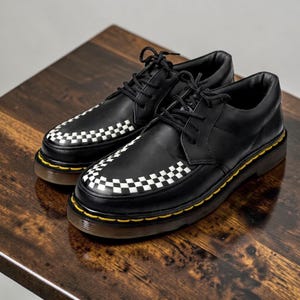 May include: A pair of black leather shoes with a white and black checkered pattern on the toe and a yellow trim. The shoes have black laces and a thick sole. The shoes are displayed on a dark brown wooden surface.