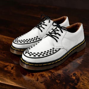 May include: A pair of white leather shoes with black laces and a checkered pattern on the toe and sides. The shoes have a brown sole with yellow stitching and are displayed on a wooden surface.