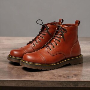 Könnte beinhalten: Ein Paar braune Lederstiefel mit Schnürung. Die Stiefel haben eine runde Spitze, eine dicke Sohle und Messingösen. Die Stiefel stehen auf einer Holzoberfläche.