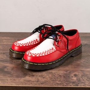 May include: Red and white leather shoes with black laces. The toe box features a white checkered pattern. The shoes have a black sole with yellow stitching and silver metal accents. The shoes are on a wooden surface.