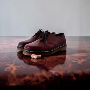 May include: A pair of burgundy leather shoes with black laces and yellow stitching around the soles. The shoes are on a reflective, brown surface. A small, white and pink object rests near the shoes.