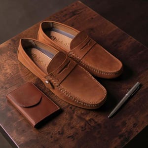 May include: A pair of brown leather loafers with a matching leather wallet and a silver pen on a wooden surface. The loafers have a classic design with visible stitching and a decorative strap. The wallet is small and rectangular.