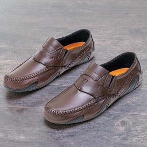 May include: A pair of brown leather slip-on shoes. The shoes have a stitched design around the toe and sides, with a dark gray sole. The interior is lined with orange fabric. The shoes are displayed on a wooden surface.