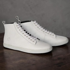 May include: A pair of high-top sneakers in solid white leather featuring matching white laces, silver-toned metal eyelets, a pull tab at the heel, and a textured white rubber sole. A small circular logo is embossed on the outer side panel.