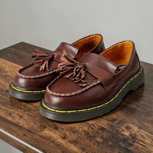 May include: A pair of brown leather loafers with decorative tassels. The shoes have a thick black sole with a yellow accent line. The interior lining is a golden yellow color. The shoes are displayed on a wooden surface.
