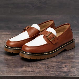 May include: A pair of two-tone brown and white leather loafers with a buckle closure. The shoes have a thick, brown sole with a black and yellow trim. The shoes are displayed on a dark wooden surface.