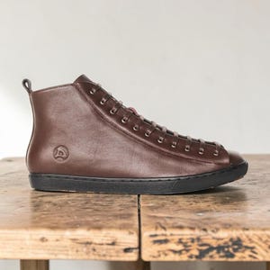 May include: A high-top sneaker in dark brown leather with a smooth finish, featuring a black rubber sole, matching dark brown laces, metal eyelets, and a small embossed circular logo on the side near the heel with a pull tab at the top.
