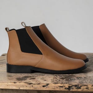 May include: A pair of tan leather Chelsea boots featuring black elastic side panels, a rounded toe, and a low black sole. Each boot includes a small pull tab at the back of the ankle collar, providing a clean and simple design for these slip-on shoes.