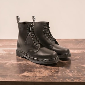 May include: A pair of black leather boots with a lace-up design. The boots have a tag attached with text and are set on a wooden surface. The boots are a classic style, suitable for various outfits.