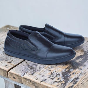 May include: A pair of black leather slip-on shoes featuring a smooth finish, rounded toes, and elastic side panels for a secure fit. The shoes have a low-profile design with matching black soles and a small embossed logo on the upper side.