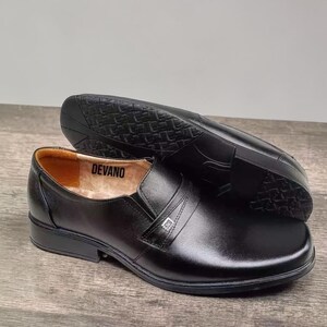 May include: Black leather slip-on shoes with a low heel. The shoes have a strap across the top with a silver-colored emblem. The brand name "DEVANO" is visible inside the shoe. The shoes are displayed on a wooden surface.
