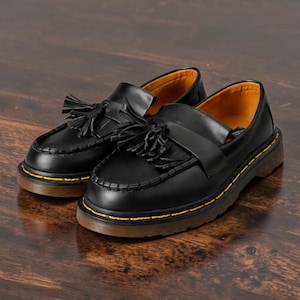 May include: A pair of black leather loafers with tassel detailing. The shoes have a rounded toe, a thick brown sole, and yellow stitching around the perimeter. The interior lining is a warm yellow color.
